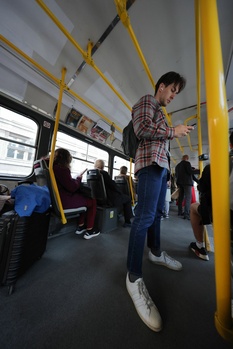 Man standing in public transit bus interior with yellow support poles and other passengers visible.