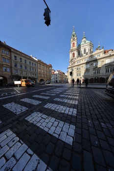 European plaza with geometric paved foreground, baroque church with twin spires in background, classical buildings flanking the square.