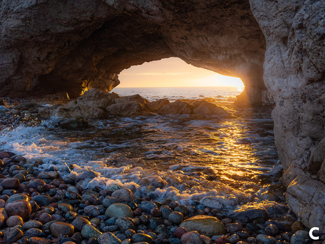 Golden sunlight streams through a sea cave opening onto a rocky beach at sunset.