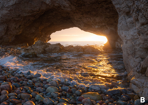 View from inside a coastal cave looking out toward golden sunset light reflecting off the ocean and rocky beach.