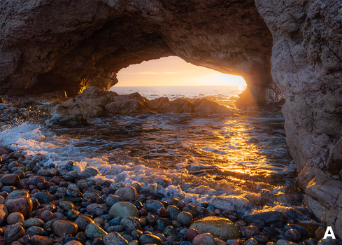 Sunlit coastal cave opening onto rocky beach and sea at golden hour.