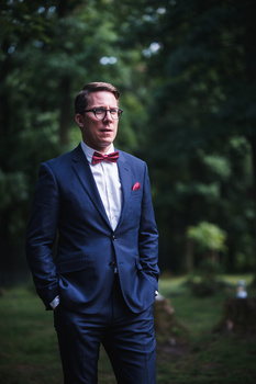 Man in navy blazer and pink bow tie standing outdoors with blurred green foliage background.