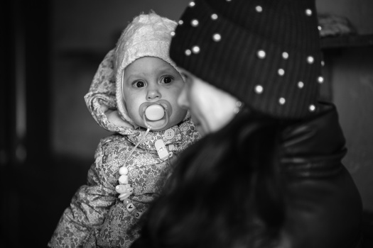 Infant with pacifier held by adult wearing black coat with pearl beads in black and white portrait.