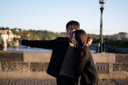 Man pointing toward distant cityscape while posing with woman against stone waterfront wall.