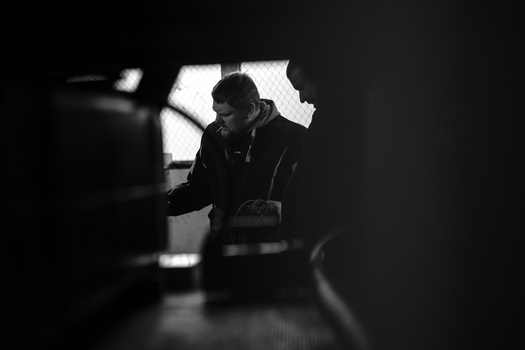 Man in dark hoodie leaning forward near bright window light in high-contrast black and white photograph.
