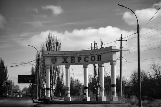 Monochrome photograph of a neoclassical gateway structure with Cyrillic signage and tall poplar trees flanking the colonnade.