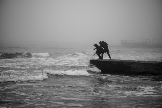 Surfer in wetsuit balancing on a wooden log in choppy ocean waves under overcast skies.