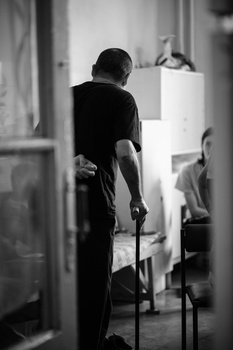 Elderly man with cane walking through a narrow hallway in black and white.