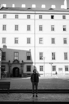 Person standing alone in a plaza before a classical European apartment building in black and white.