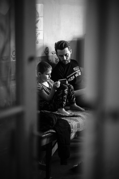 Man teaching young boy to play acoustic guitar in a narrow interior space, shot through a foreground obstruction.