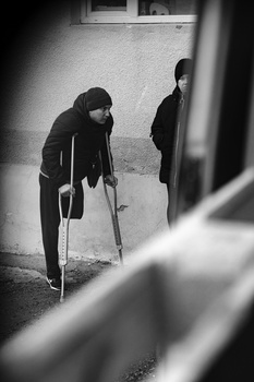 Two figures in dark clothing on a street, one in foreground with crutches, viewed through blurred architectural elements.