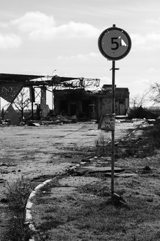 Rusted speed limit sign reading 5 in an abandoned industrial area with dilapidated structures.