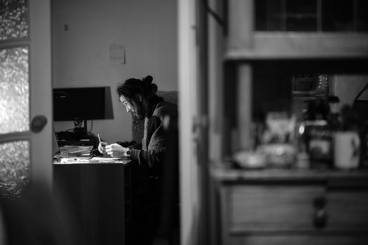 Person working at a desk in a darkened room, photographed through a doorway in black and white.