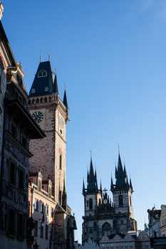 Gothic church spires and a historic stone tower rise against a clear blue sky in an old European city center.