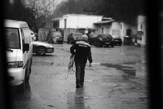 Person in dark clothing walking away across wet pavement toward parked vehicles in a blurred urban lot.