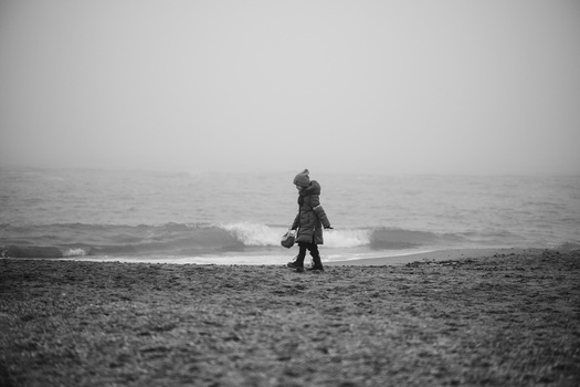 Child standing alone on a pebbly beach facing the ocean on an overcast day.