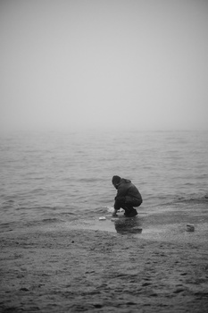 Solitary figure crouched on a misty beach, rendered in black and white photography.