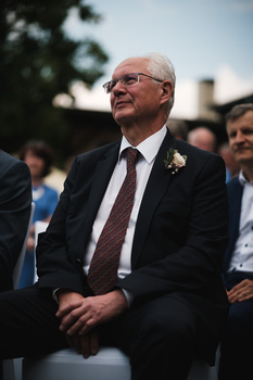 Older man in dark suit and burgundy tie with boutonniere, smiling while looking off-camera at an outdoor event.