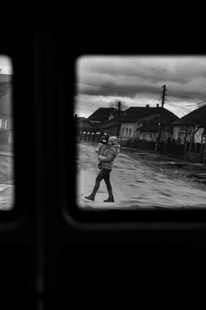 Black and white photograph framed through a window, showing a solitary figure walking on a wet street with buildings and overcast sky.
