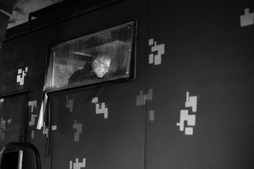 Black and white image of a person visible on a mounted monitor against a dark wall decorated with white cross shapes.