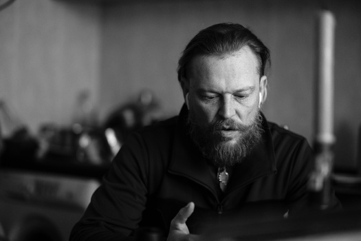 Bearded man in dark clothing looking downward in a contemplative pose.