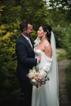 Bride and groom face each other outdoors, holding a bouquet, surrounded by green foliage.