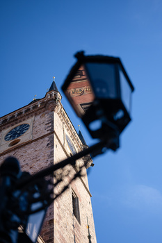 Historic stone clock tower shot from below against blue sky, with a street lamp in sharp focus in the foreground.