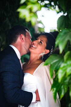 Bride and groom embracing and smiling outdoors, framed by green foliage.