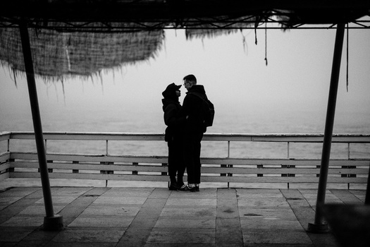 Two silhouetted figures standing close together on a covered waterfront deck, framed by wooden posts and railings.