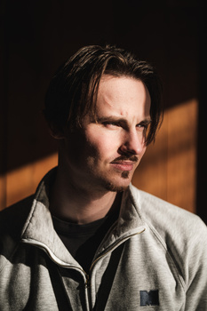 Man with dark hair and facial hair wearing a light gray jacket over dark clothing, photographed against a dark background with warm side lighting.