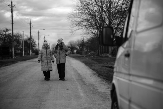 Two figures in winter coats walking away down a rural road lined with bare trees and utility poles.