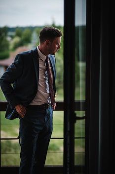Man in dark blazer and tie standing by large window with green landscape view.