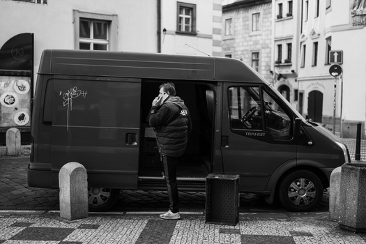 Person standing in open doorway of dark van parked on European street lined with bollards.
