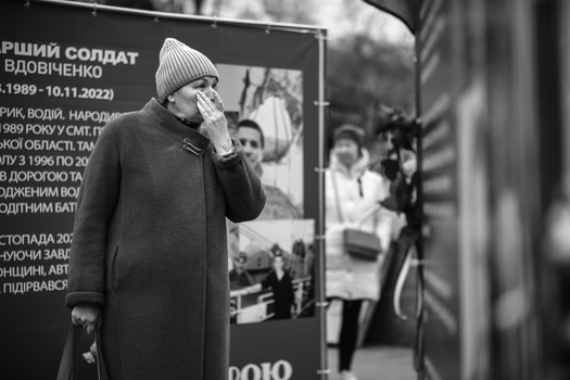 Person in winter coat and beanie standing before a memorial plaque, hand to face in gesture of grief.