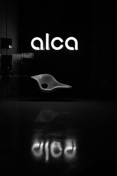 Minimalist product photograph of a curved white sculptural form displayed against a dark background with its reflection visible below.