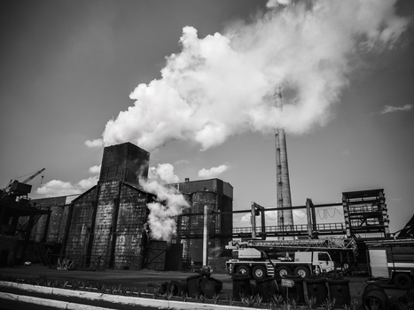 Industrial facility with grain elevators, smokestacks, and trucks under a cloudy sky in black and white.