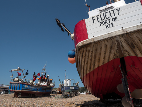 Fishing vessel with red and white hull beached on shingle, with blue fishing boat and industrial structures visible in background.