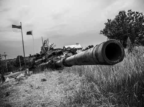 Rusted tank barrel in foreground with abandoned military wreckage and flags on hillside behind it.