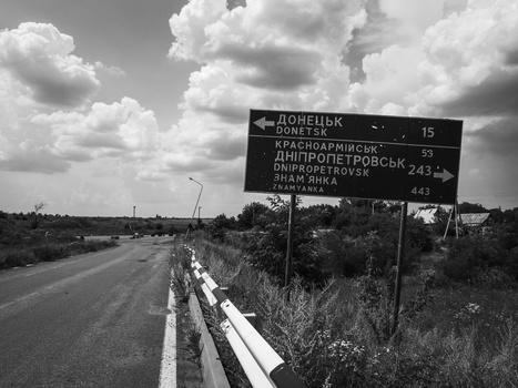 Road sign with Cyrillic text indicating directions to cities, captured in black and white with dramatic cloudy sky.