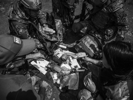 Overhead view of multiple hands in camouflage clothing gathered around scattered papers and documents on the ground.