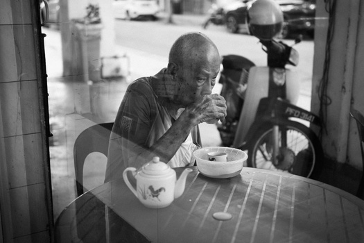 Man in dark shirt seated at table with tea service, photographed through window with street scene visible behind.