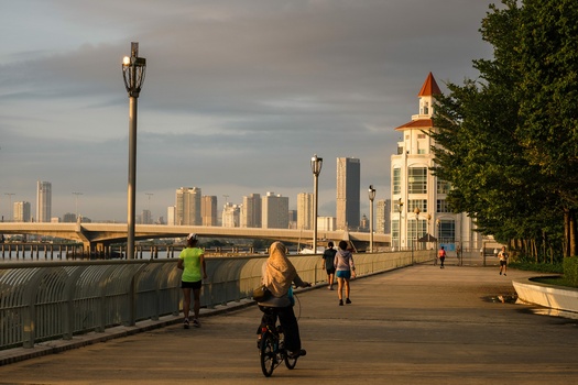 People cycling and walking on a waterfront promenade with a city skyline across the water at golden hour.