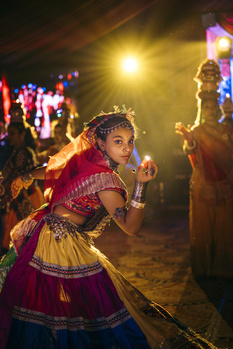 Performer in ornate traditional costume dancing at night festival with colorful lights in background.