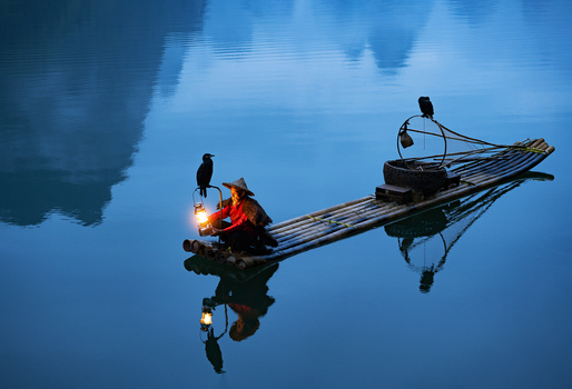 Two traditional wooden boats with cormorant birds and lanterns reflected in still blue water.
