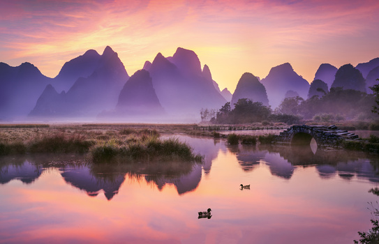 Misty karst mountains reflected in still water at dawn, with small boats and riverside vegetation.