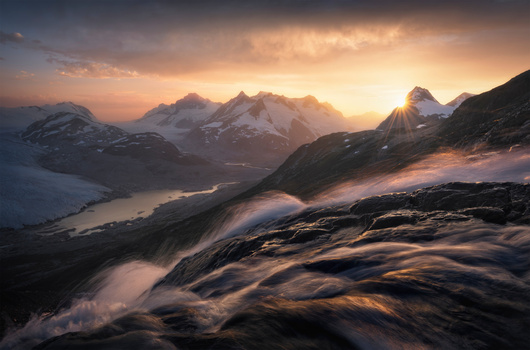 Snow-covered mountain range at sunset with glacial valley and flowing mist in foreground.