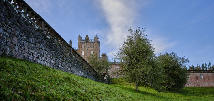 Stone castle with multiple turrets set on a hillside surrounded by green grass and trees under partly cloudy sky.