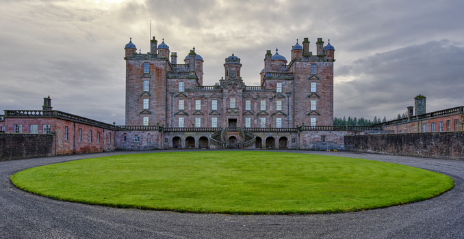 Red stone castle with multiple towers surrounding a circular green lawn in symmetrical composition.