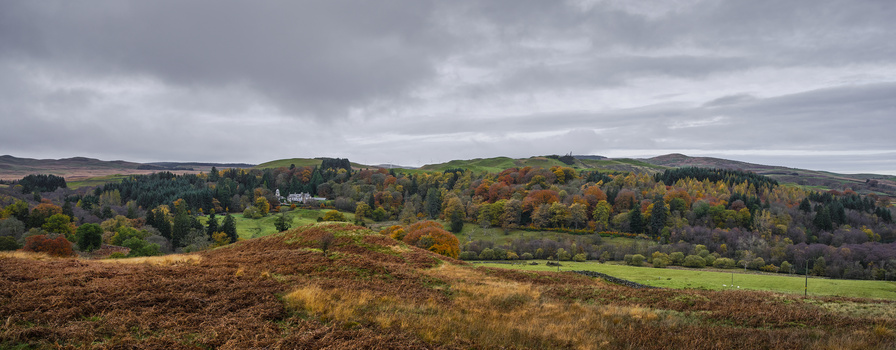 Rolling moorland and forested valley under overcast sky with distant mountains.
