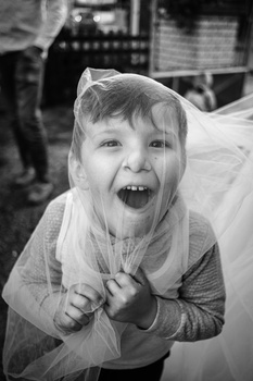 Young child wearing a sheer veil, smiling with joy and excitement at an outdoor event.